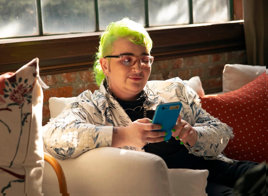 A genderqueer person with neon hair sitting on a couch looking at their phone.